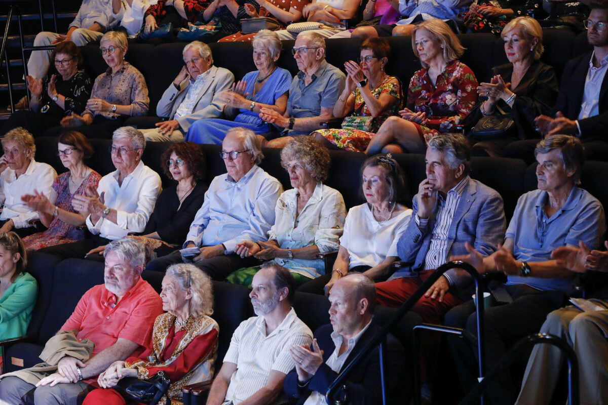 A group of older adults sit closely together in theatre-style rows, attentively watching and applauding an event. Most are dressed in smart casual or formal clothing. The mood appears engaged and appreciative.