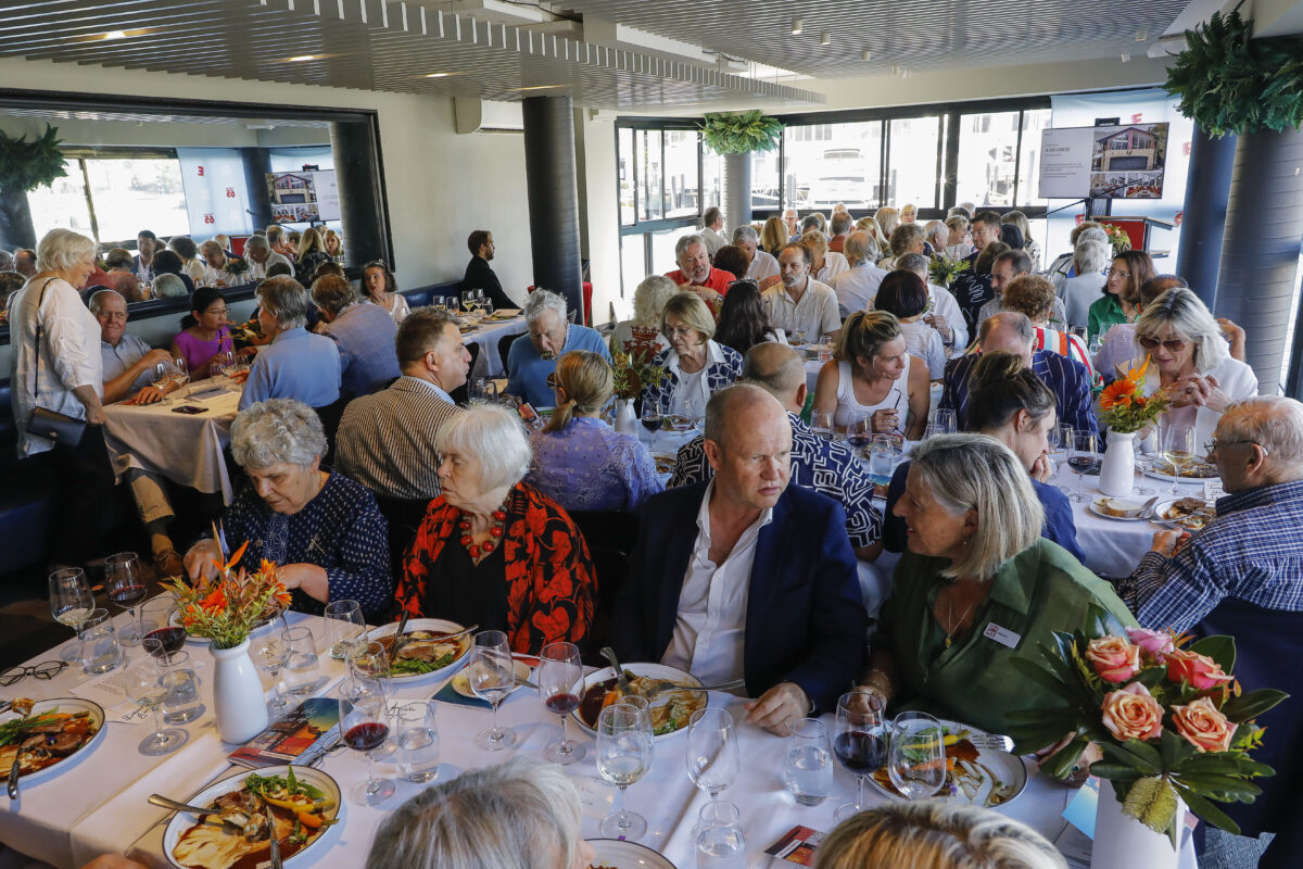 A large group of people gather around several tables set for a meal, engaged in conversation and dining. The atmosphere is lively, with flower centrepieces and glasses on the tables in a brightly lit room.