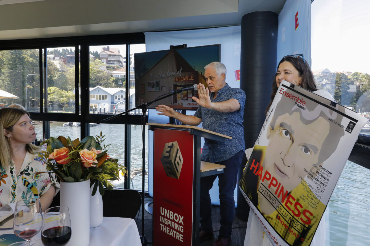 A man speaks at a lectern beside a woman holding a large theatre poster titled Happiness, while attendees sit at a table with flowers and wine in a room overlooking water through large windows.