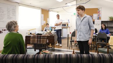 Five people are in a casual office or rehearsal space. One young man stands and speaks to an older woman seated on a sofa, while three others watch, sitting or standing in the background. The atmosphere appears serious.