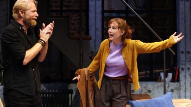 A man with a beard, wearing a black shirt, claps his hands and smiles at a woman in a yellow cardigan who gestures animatedly, standing in a modern interior with stairs and exposed brick in the background.