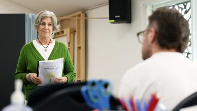 An older woman in a green jumper holds a script and stands at the front of a classroom, facing a seated man. Classroom supplies and a window are visible in the background.