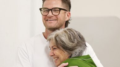 A man wearing glasses and a white shirt hugs an older woman in a green jumper. Both are smiling, and the embrace appears warm and joyful. The background is plain and light-coloured.