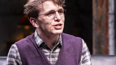 A young man with short brown hair, glasses, and a purple jumper vest over a checked shirt sits on a black sofa, speaking with an expressive face. Warm light and a blurred, cosy background set the scene.