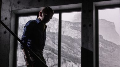 A man in a dark shirt and trousers walks down an indoor staircase beside large windows, with light streaming in and mountains visible outside. The scene is shadowy and dramatic.