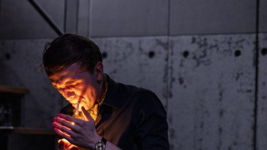 A man in a dark shirt stands indoors by a concrete wall, lighting a cigarette. The glow from the lighter illuminates his face and hands. There are stairs and large windows showing mountains in the background.