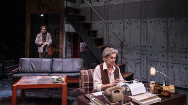 An older woman sits at a desk typing on a typewriter in a modern living room. Papers, books, and a phone clutter the desk. A younger man stands in the background near a staircase, watching her. The scene suggests a tense moment.