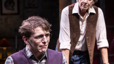 A young man with glasses and a purple jumper sits tensely on a sofa, hands clasped, while an older woman in a waistcoat leans forward from behind, watching him with concern. The setting appears to be a dimly lit living room.