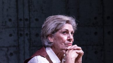 An older woman with grey hair sits thoughtfully at a desk, hands clasped under her chin, in front of a typewriter. She wears a white shirt and brown waistcoat, with a checked blanket draped over the chair.
