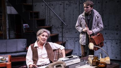 An older woman sits at a cluttered desk with a typewriter, talking to a younger man standing nearby with a satchel, set in a dimly lit, industrial-style office or study.