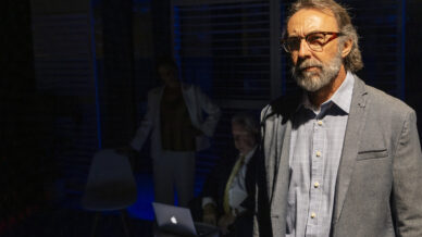 A man with grey hair, beard, and glasses stands in the foreground wearing a blazer. Behind him, two people sit by a laptop in a dimly lit room with blue light.
