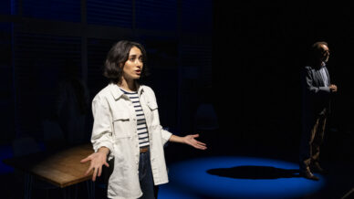 A woman stands in a spotlight with her arms open, appearing to speak or perform. She wears a light jacket over a striped shirt. In the background, a man stands in a separate spotlight, both on a dark stage.