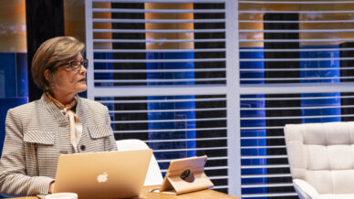 A woman wearing glasses and a suit sits at a desk with a laptop, tablet, and coffee mug. Behind her are large windows with blinds, revealing a cityscape at sunset.