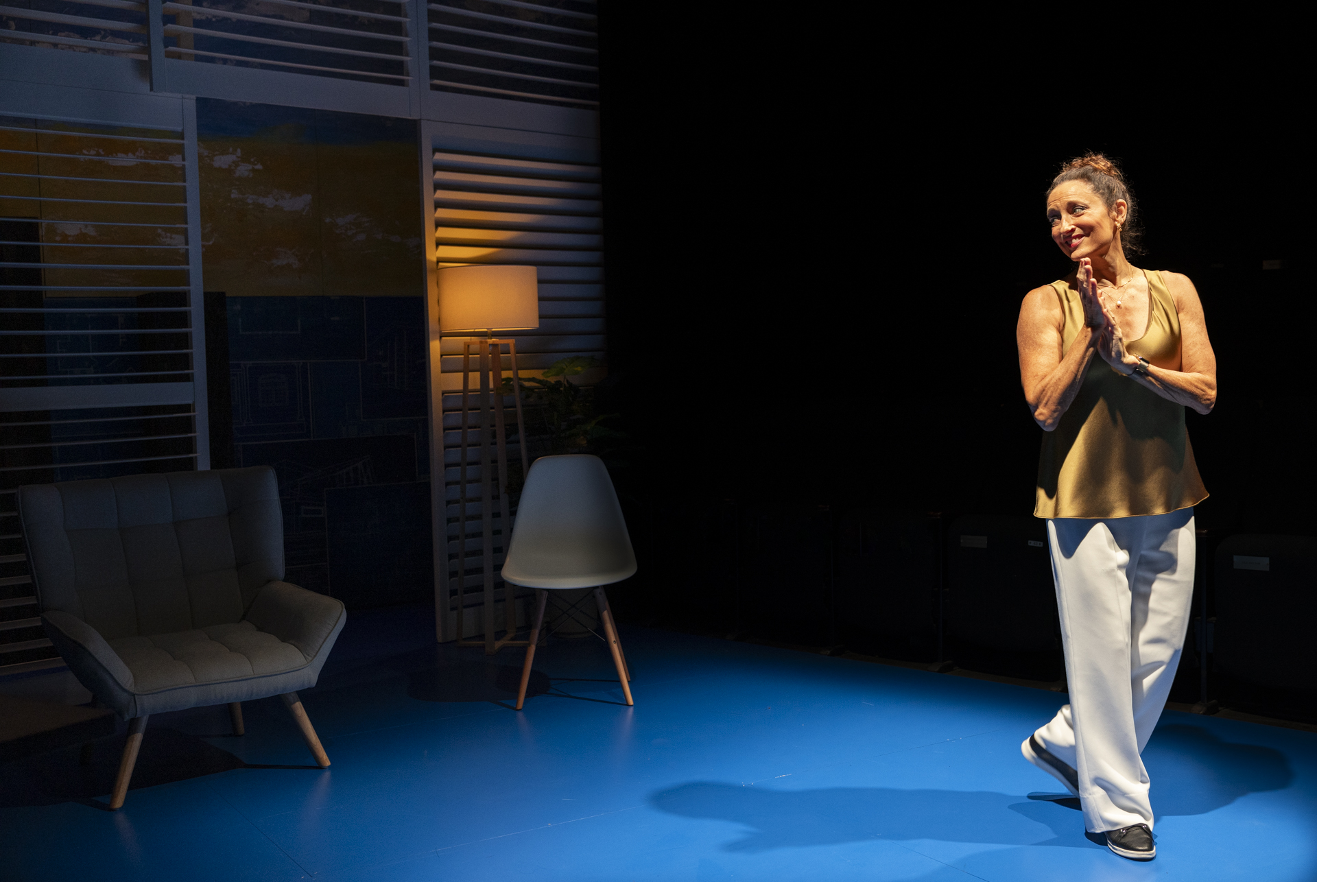 A woman stands on a brightly lit blue stage, smiling with her hands clasped. She wears a tan top and white trousers. Behind her are modern chairs, a standard lamp, and window blinds, with the background partially in shadow.