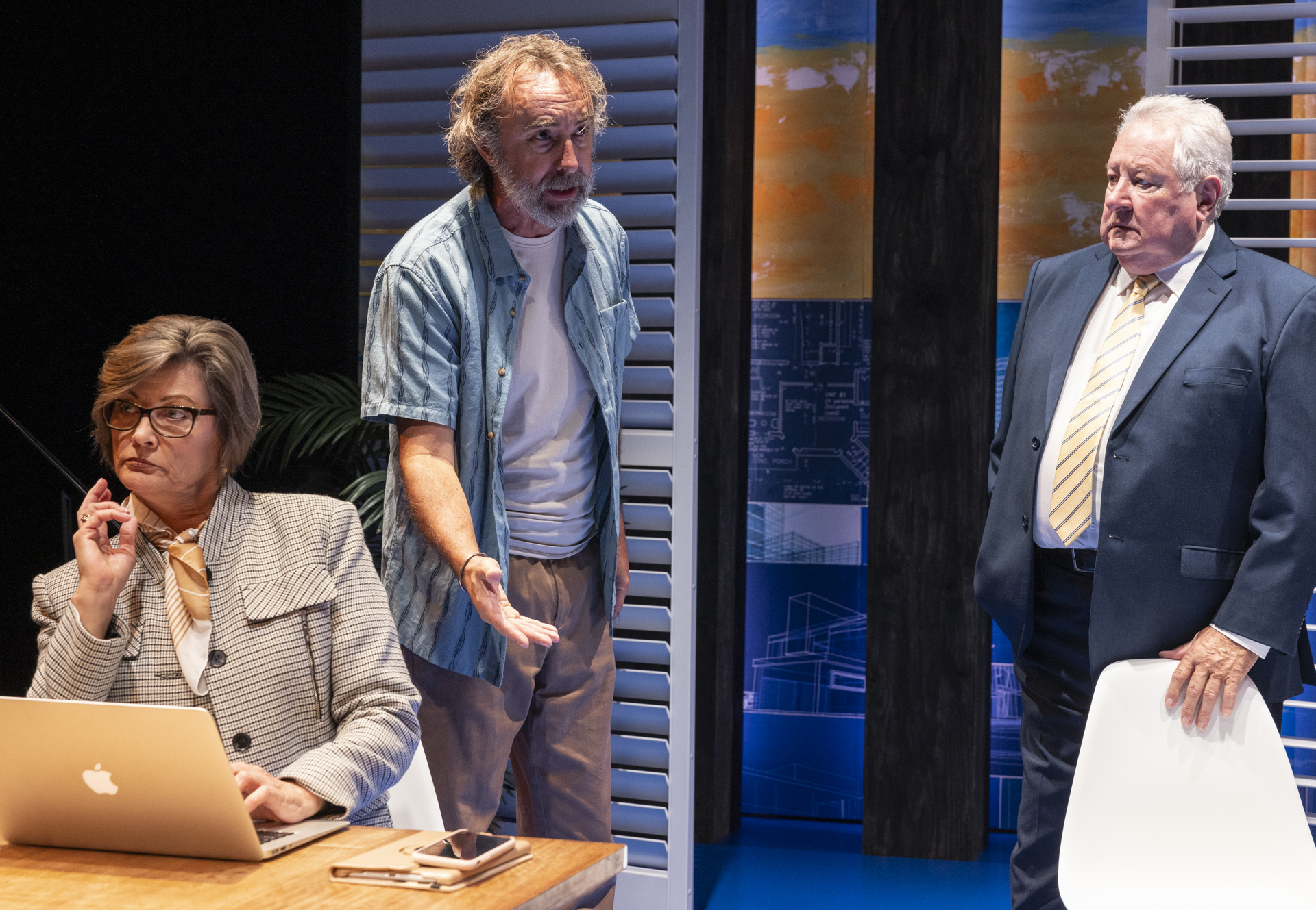 A woman in a suit sits at a desk with a laptop, looking thoughtful. An older man in casual clothes gestures as he speaks, whilst another man in a suit stands nearby, watching seriously.