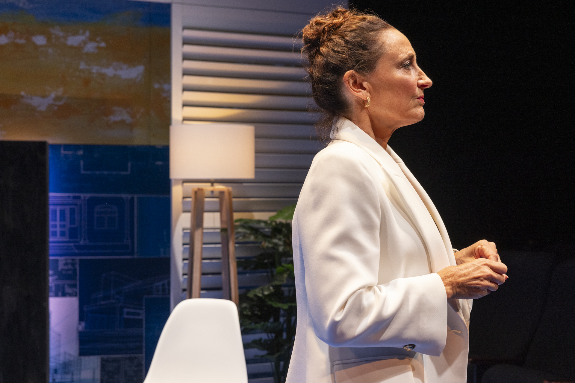 A woman with brown hair in a bun, wearing a white blazer, stands in profile indoors. Behind her are a white chair, standard lamp, green plant, and colourful wall art. The setting appears modern and stylish.