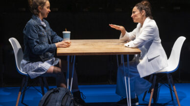 Two women sit opposite each other at a wooden table on stage. One, in a denim jacket, holds a drinks cup; the other, in a white suit, gestures while speaking. A rucksack rests near the table on a blue floor.