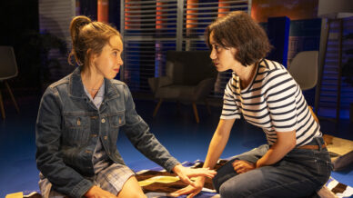 Two women sit on a striped blanket indoors, facing each other. One wears a denim jacket and dress, the other wears a striped shirt and jeans. They appear to be having an intense, emotional conversation, with warm lighting around them.