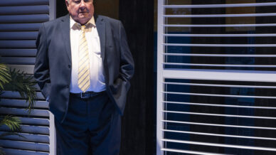 An older man in a dark suit and yellow-striped tie stands with his hands in his pockets beside large window blinds. A potted plant is nearby, and blue lighting creates a formal, indoor atmosphere.