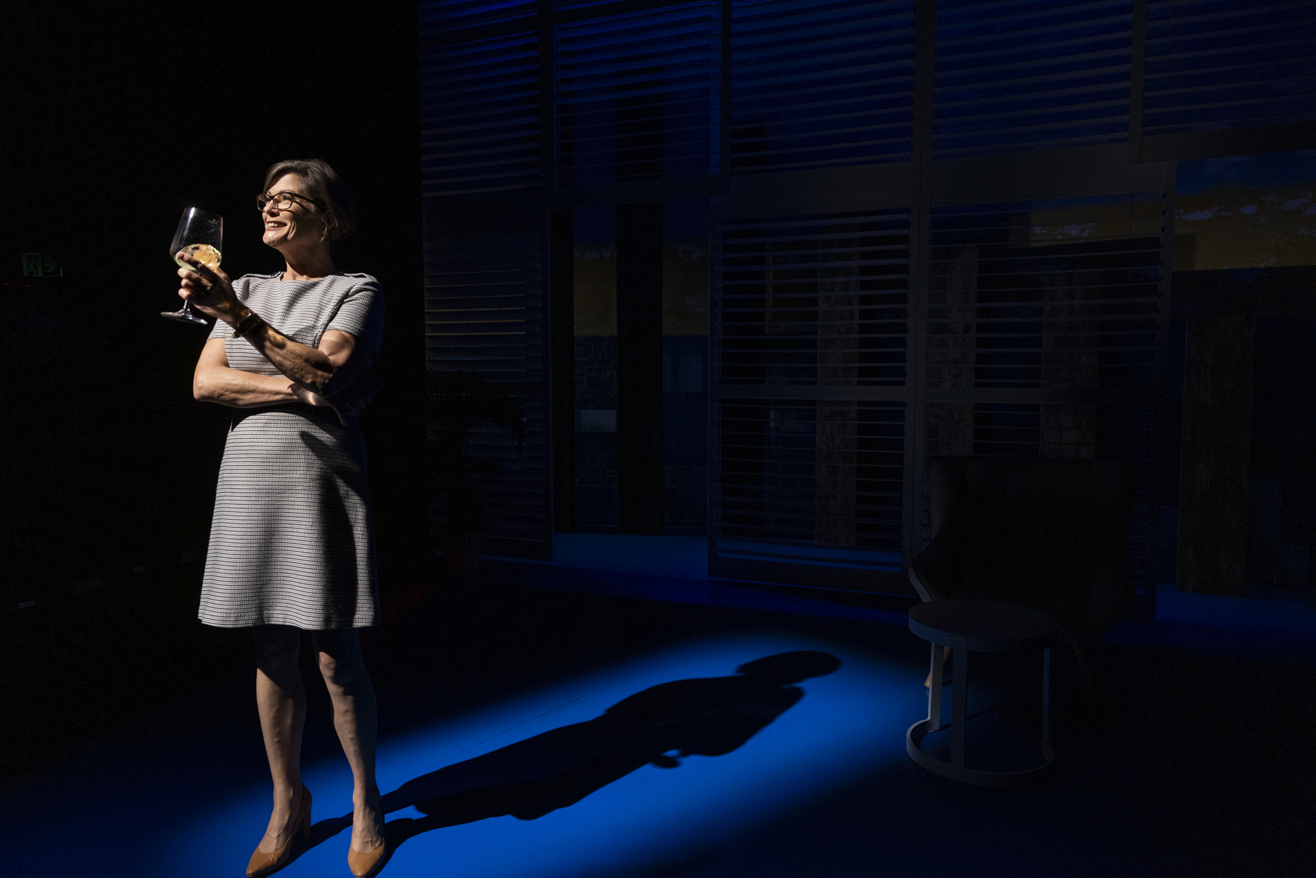 A woman in a grey dress stands under a spotlight on a dark stage, smiling whilst holding a wine glass and a pastry. Her shadow is cast on the blue-lit floor behind her.