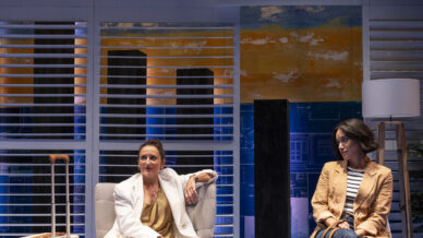 Two women sit on stage in a modern set. One, in a white suit, sits confidently in an armchair next to a suitcase. The other, dressed casually in jeans and a tan jacket, sits on a bench with a large bag, looking away.