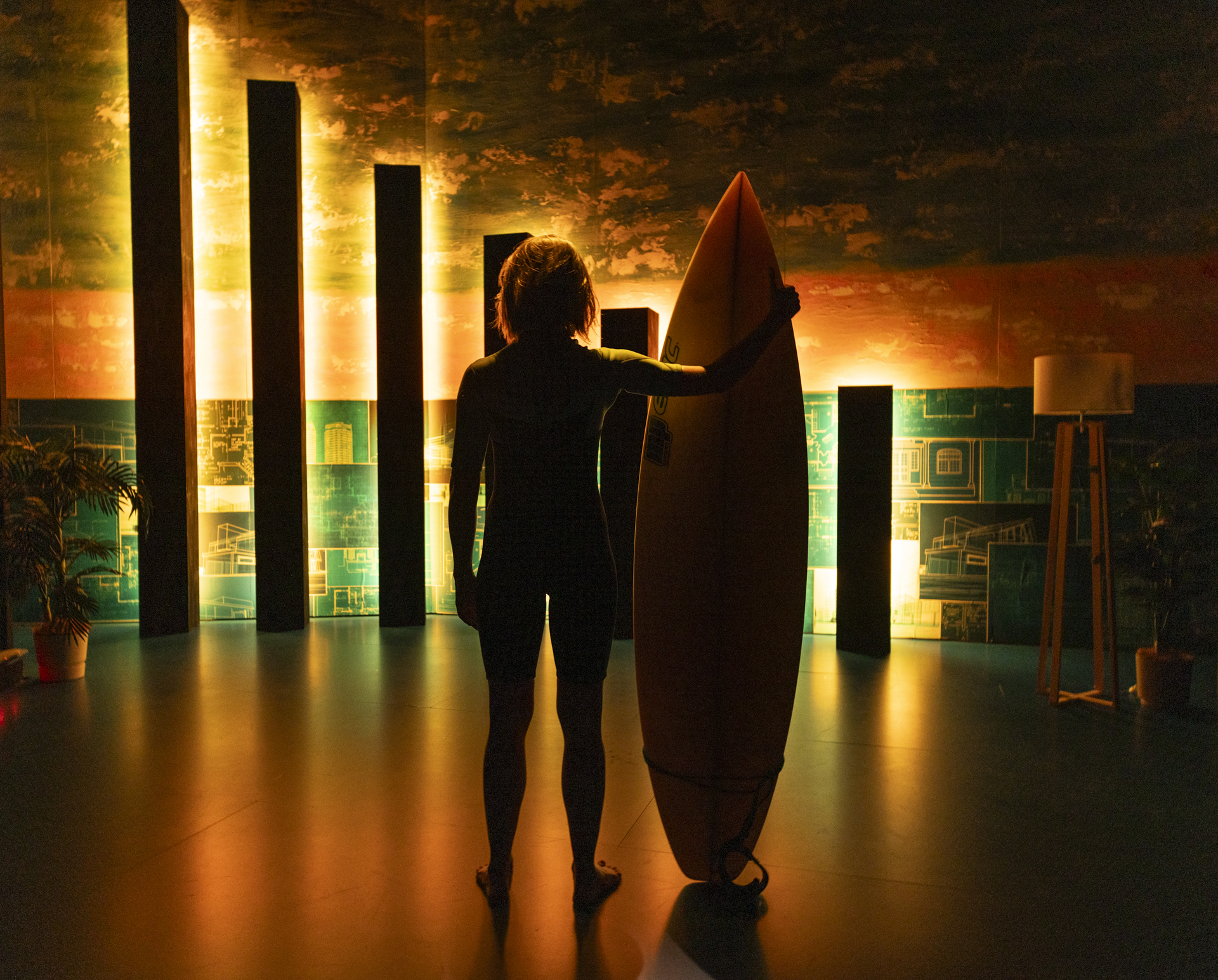 A person stands indoors holding a surfboard, silhouetted against a warm, glowing background with vertical light panels and decorative plants.