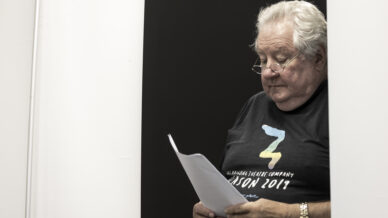 An older man with white hair and glasses stands reading from a script. He wears a black T-shirt with colourful text and a watch, standing against a stark black and white background.