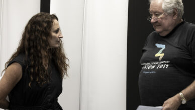A woman with long curly hair in a dark dress faces an older man with grey hair and glasses, who is holding papers and wearing a black T-shirt with colourful text. They appear to be having a serious conversation indoors.