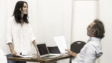A woman stands and smiles whilst talking to a seated man holding a paper. They are at a table with a laptop and documents in a bright room with white walls and chairs in the background.