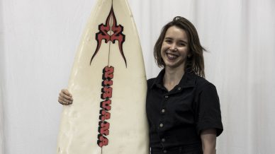 A smiling person in a black playsuit and white sandals stands indoors, holding a white surfboard with red and black designs, in front of a white curtain background.