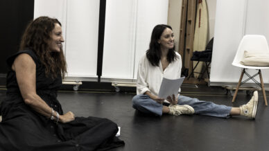Two women sit on the floor indoors, smiling and holding papers. One wears a black dress, the other a white shirt and jeans with trainers. Behind them are white panels, a chair, and a surfboard in the background.