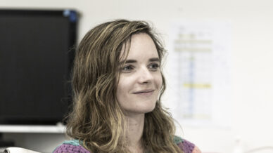 A woman with wavy hair wearing a colourful, striped jumper sits at a desk and smiles. Office equipment and documents are visible in the background.