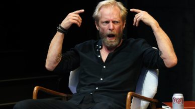 A bearded man in a black shirt sits in a chair, raising both hands and pointing at his head with an expressive, open-mouthed look. There is a can of Coca-Cola on the table next to him.