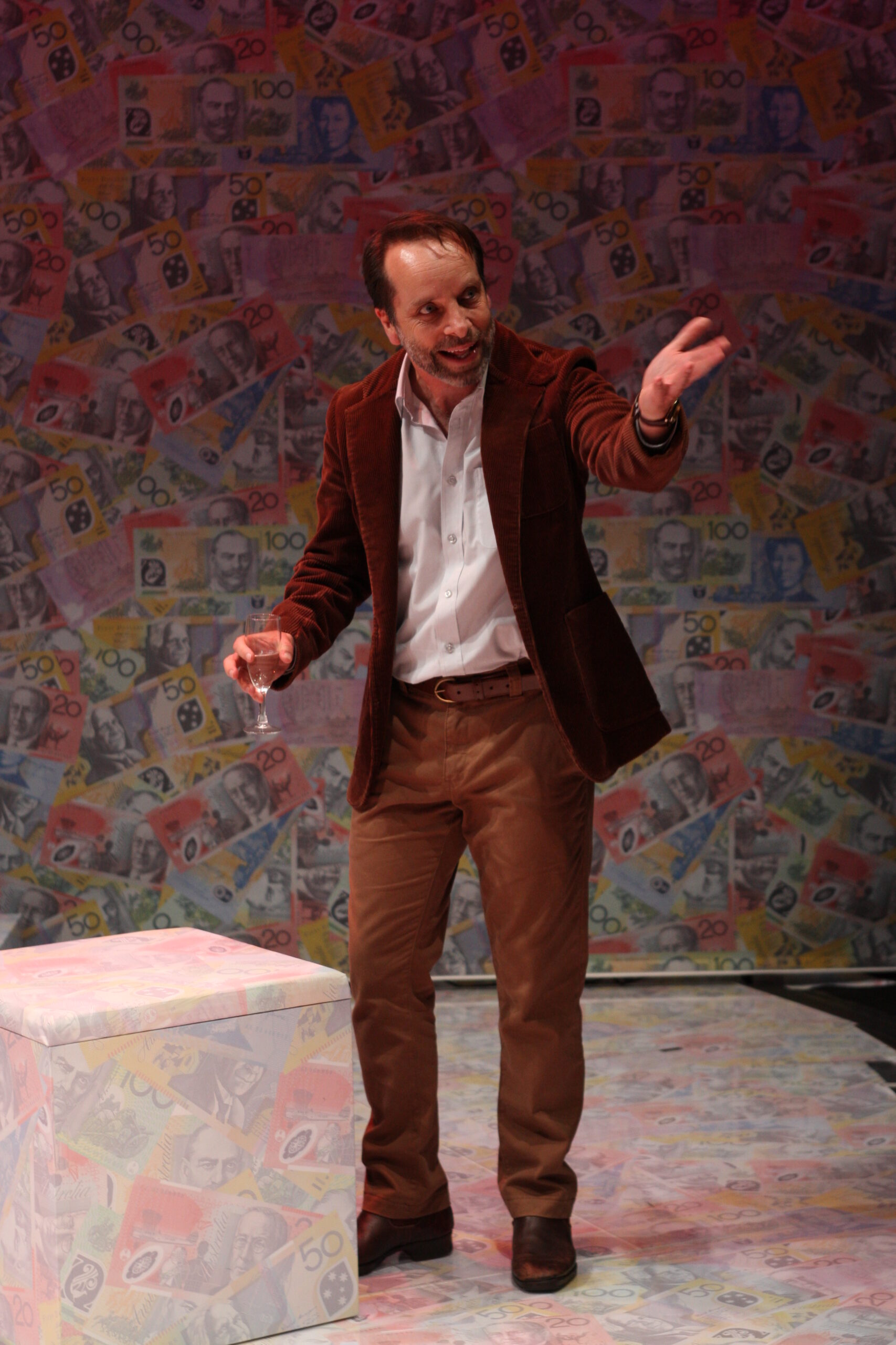 A man in a brown blazer and tan trousers stands with a drink in one hand, gesturing with the other, in a room covered with colourful banknotes.