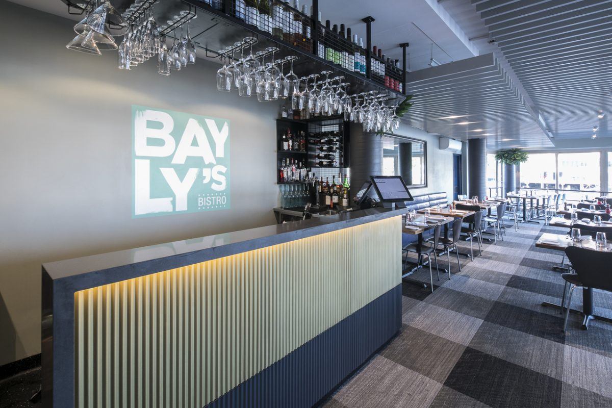 Modern restaurant interior with a sleek bar counter, hanging glasses, shelves with wine bottles, and a sign on the wall reading BAYLYS BISTRO. Dining tables and chairs are arranged neatly in the background.