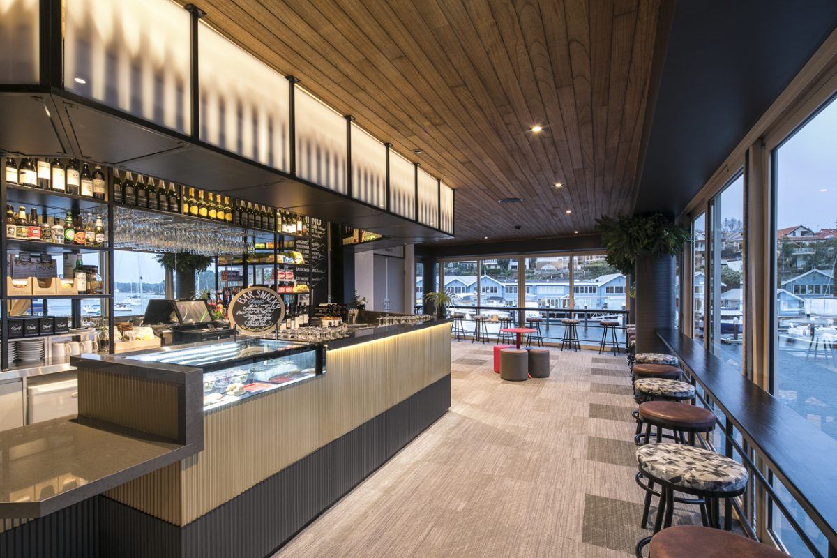 Modern café with a wooden ceiling, large windows overlooking a marina, a display counter with pastries, bar seating along the windows, and shelves with bottles and glasses behind the counter.