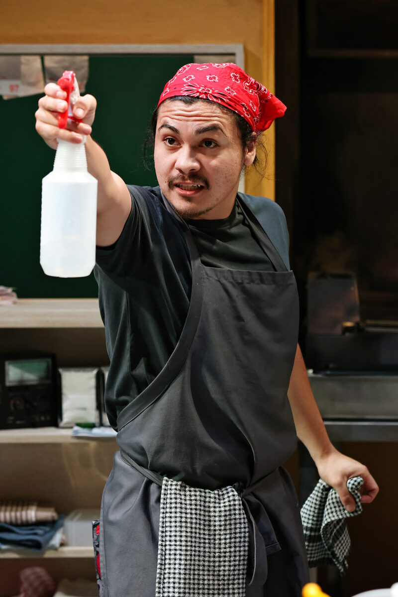 A man wearing a red bandana and dark apron holds up a spray bottle, pointing it forward. He has a determined expression and clutches a checked towel in his other hand while standing in a kitchen setting.