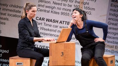 Two women are onstage; one sits at a wooden box with a laptop and looks serious, while the other, wearing dungarees, leans on another box and smiles. Large classified adverts are displayed on the backdrop behind them.
