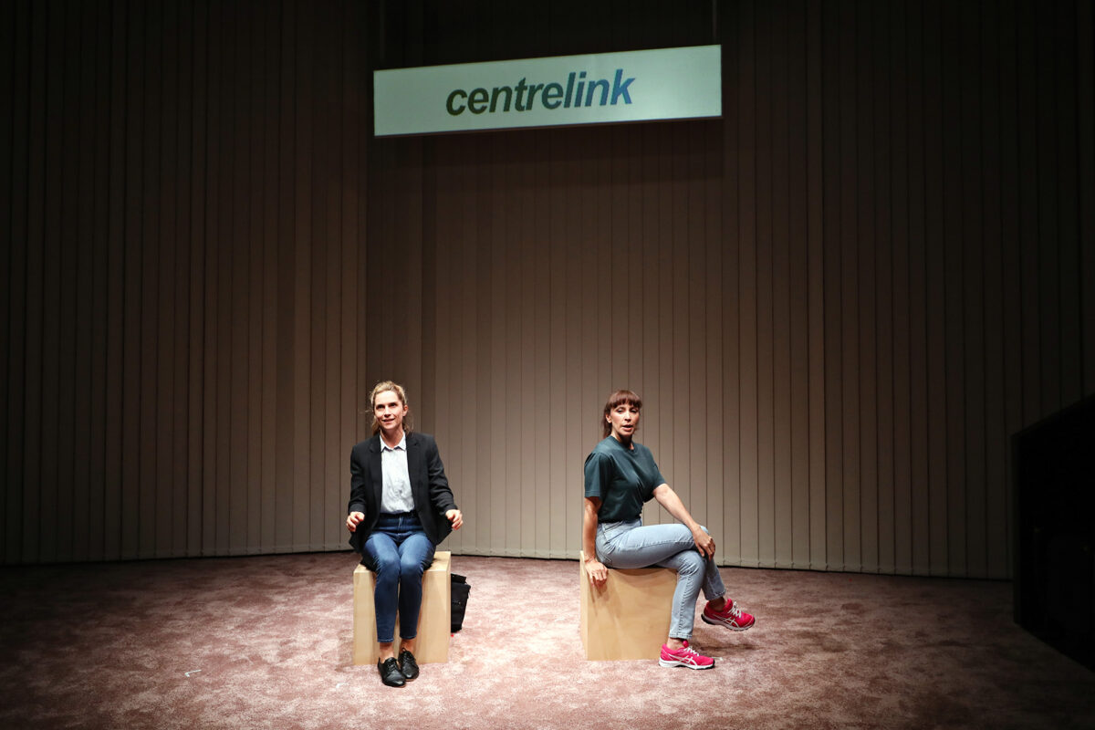 Two people sit on separate wooden boxes under a sign that reads centrelink in a sparsely furnished, carpeted room with vertical blinds in the background.