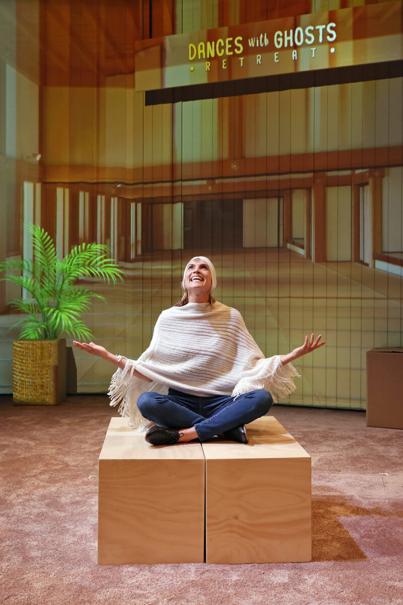 A woman sits cross-legged on a wooden platform, smiling with arms open, beneath a sign that reads DANCES with GHOSTS RETREAT in a warmly lit, modern room with a plant in the background.