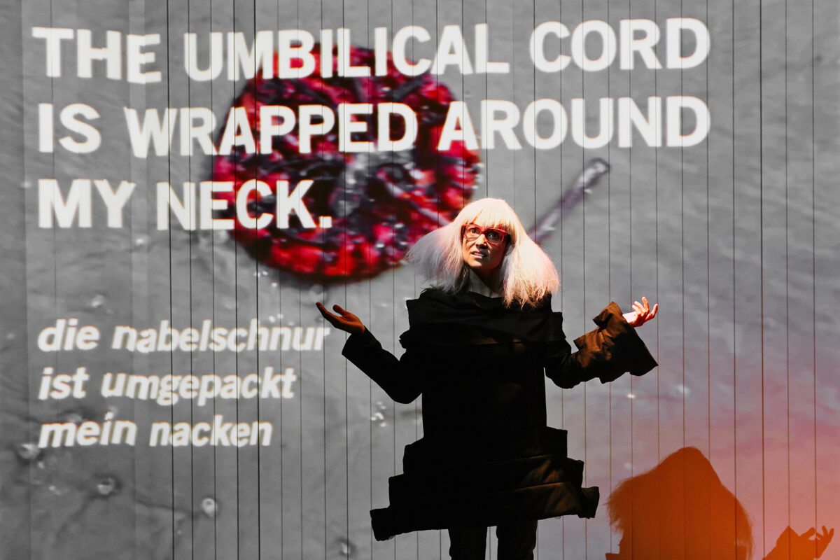 A person with white hair and glasses stands with arms outstretched in front of a screen displaying the text “THE UMBILICAL CORD IS WRAPPED AROUND MY NECK” and its German translation. A red, circular object is projected behind them.