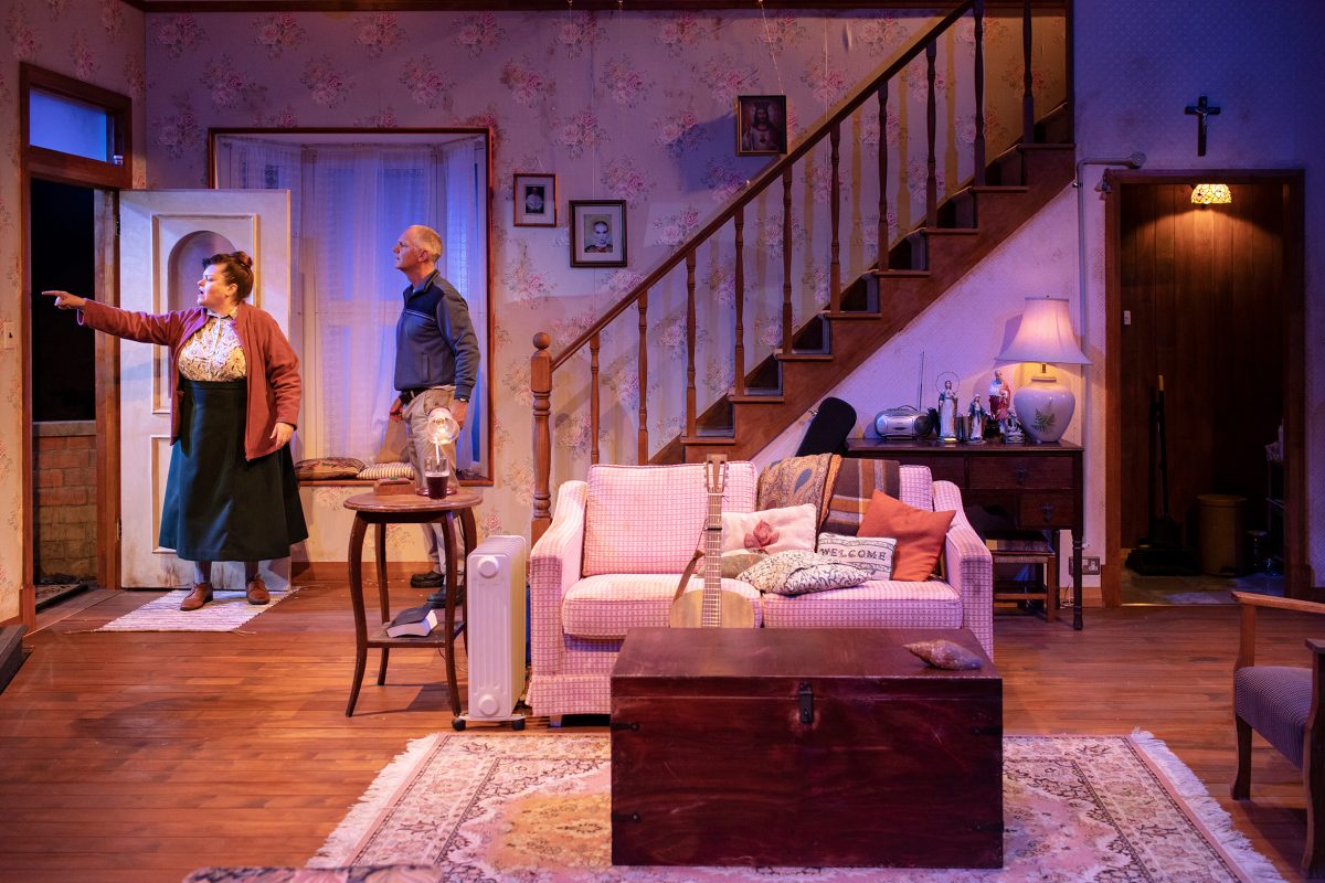 A woman points outside whilst a man stands beside her in a cosy, vintage-style living room with floral wallpaper, a staircase, and a pink sofa with cushions. Warm lighting creates a homely atmosphere.