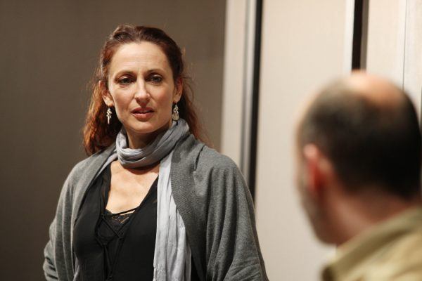 A woman with long brown hair, wearing a grey scarf and dark top, looks thoughtfully at a man in the foreground whose face is out of focus. They appear to be engaged in a conversation indoors.