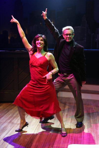 A woman in a red dress and a man in a dark suit pose energetically on a stage, each raising one arm and smiling, with a piano and cityscape backdrop behind them.
