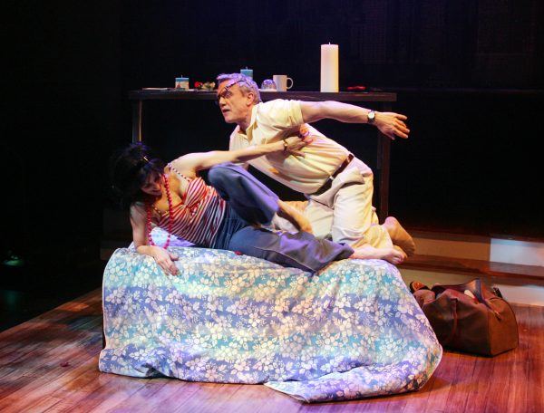 A man and woman struggle on a bed covered with a blue floral duvet. The man, wearing glasses and light clothing, grabs the woman, who is in a striped top, as they appear to fall off the bed on a wooden stage.