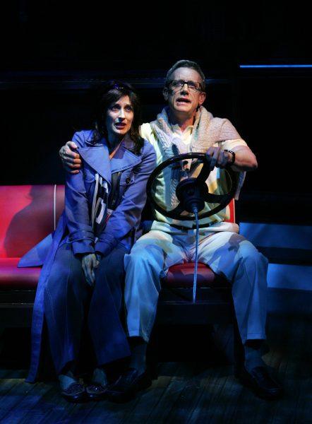 Two people sit close together on a bench, acting as if they are driving. The man holds a steering wheel and wears glasses, a yellow shirt, and a jumper over his shoulders. The woman wears a blue coat. Stage lighting creates a dramatic effect.