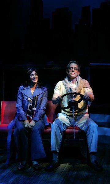Two actors on stage simulate sitting in a car; the man on the right holds a steering wheel and smiles, while the woman on the left sits beside him, looking at him. The city skyline is silhouetted in the background.