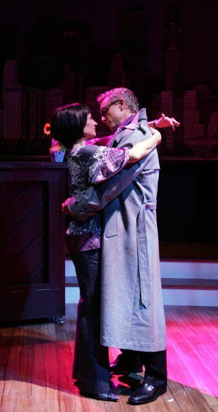 A man in a grey trench coat and glasses dances closely with a woman in a patterned blouse on a stage with city skyline backdrop and a piano nearby, both gazing at each other.