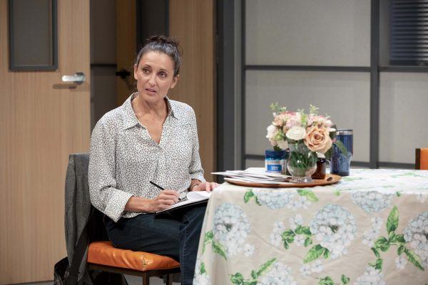A woman with dark hair tied up sits at a table covered with a floral tablecloth, holding a pen and notebook. She looks to the side with a thoughtful expression. There is a flower arrangement and papers on the table.