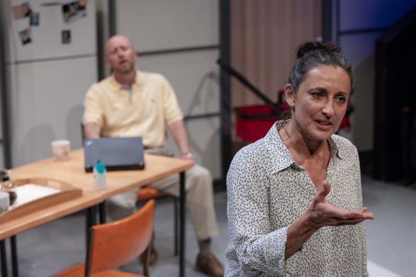 A woman gestures while speaking in the foreground, wearing a patterned blouse. In the background, a man sits at a table with a laptop, looking on. The setting appears to be a kitchen or dining area.